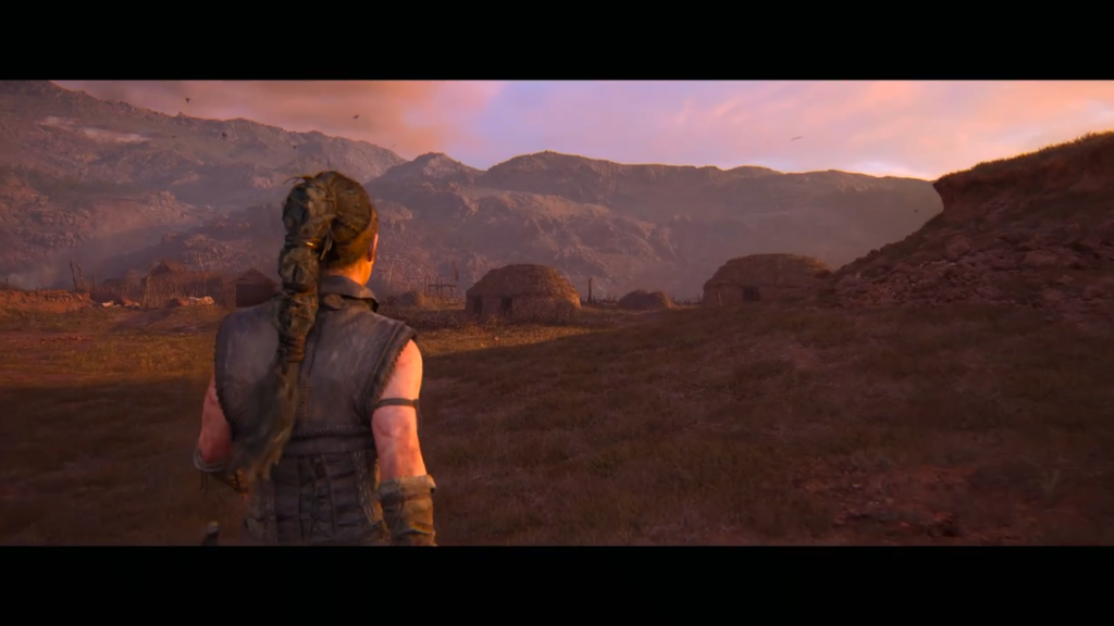 Senua walks toward huts in mountainous valley at sunset