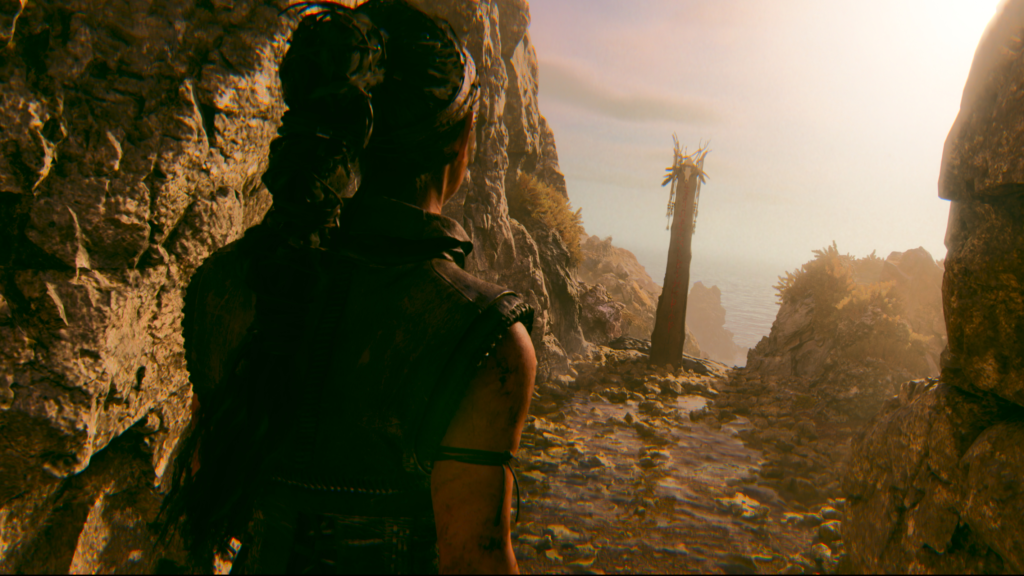 Senua walks along rocky cliff path toward sea